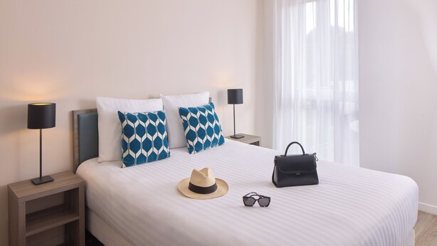 A neatly made bed with patterned pillows, a hat, sunglasses, and a handbag on the bed. - Quality Aparthotel Morangis Orly (Morangis)