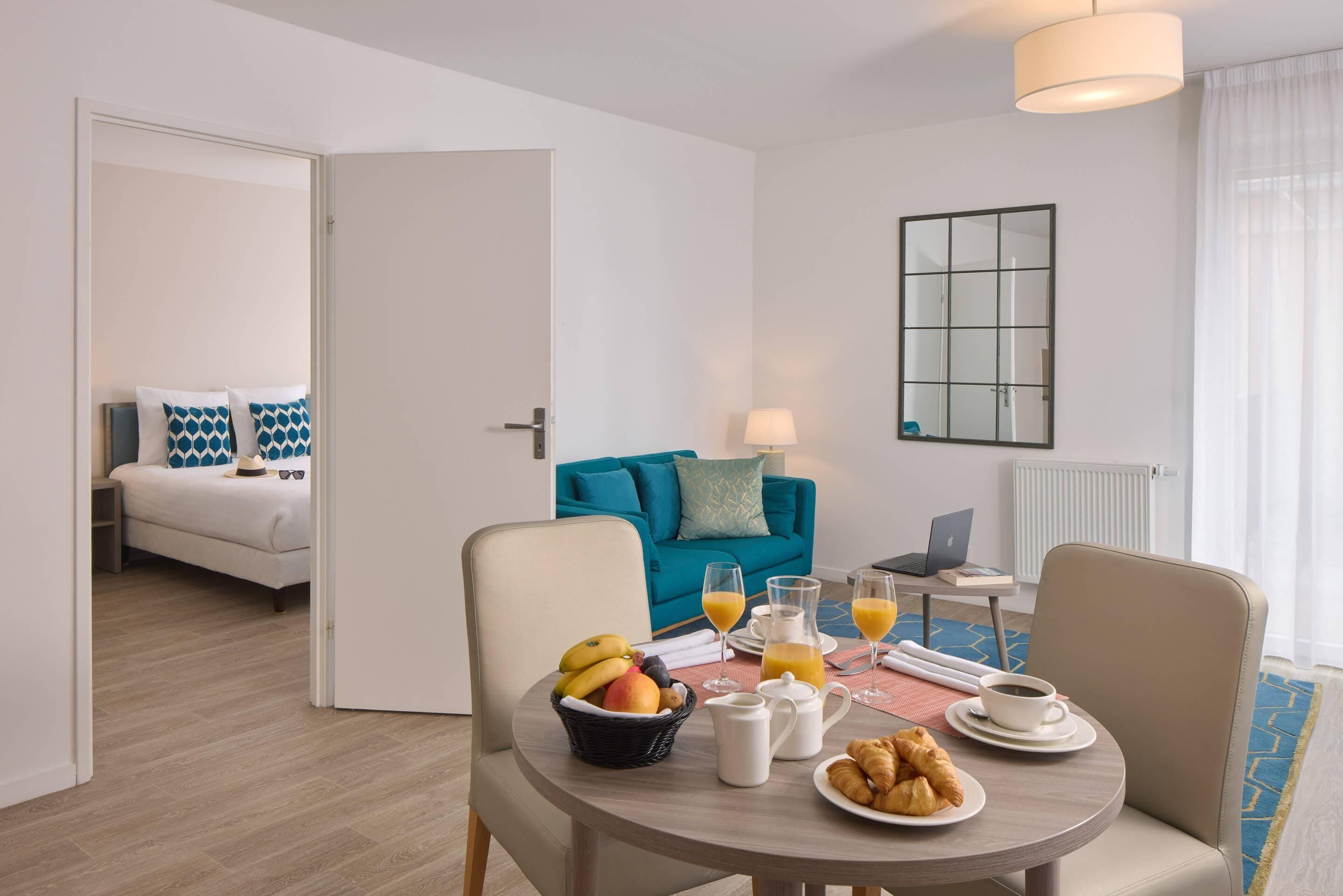 A modern hotel room with a dining area, a bed, a blue sofa, and a laptop on a table with breakfast items.