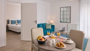 A modern hotel room with a dining area, a bed, a blue sofa, and a laptop on a table with breakfast items. - Quality Aparthotel Morangis Orly (Morangis)