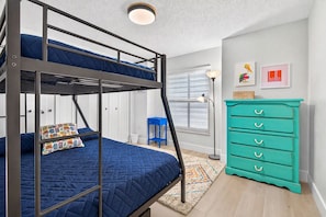 Una cama litera con colcha azul, un aparador color turquesa y una mesita de noche con lámpara.