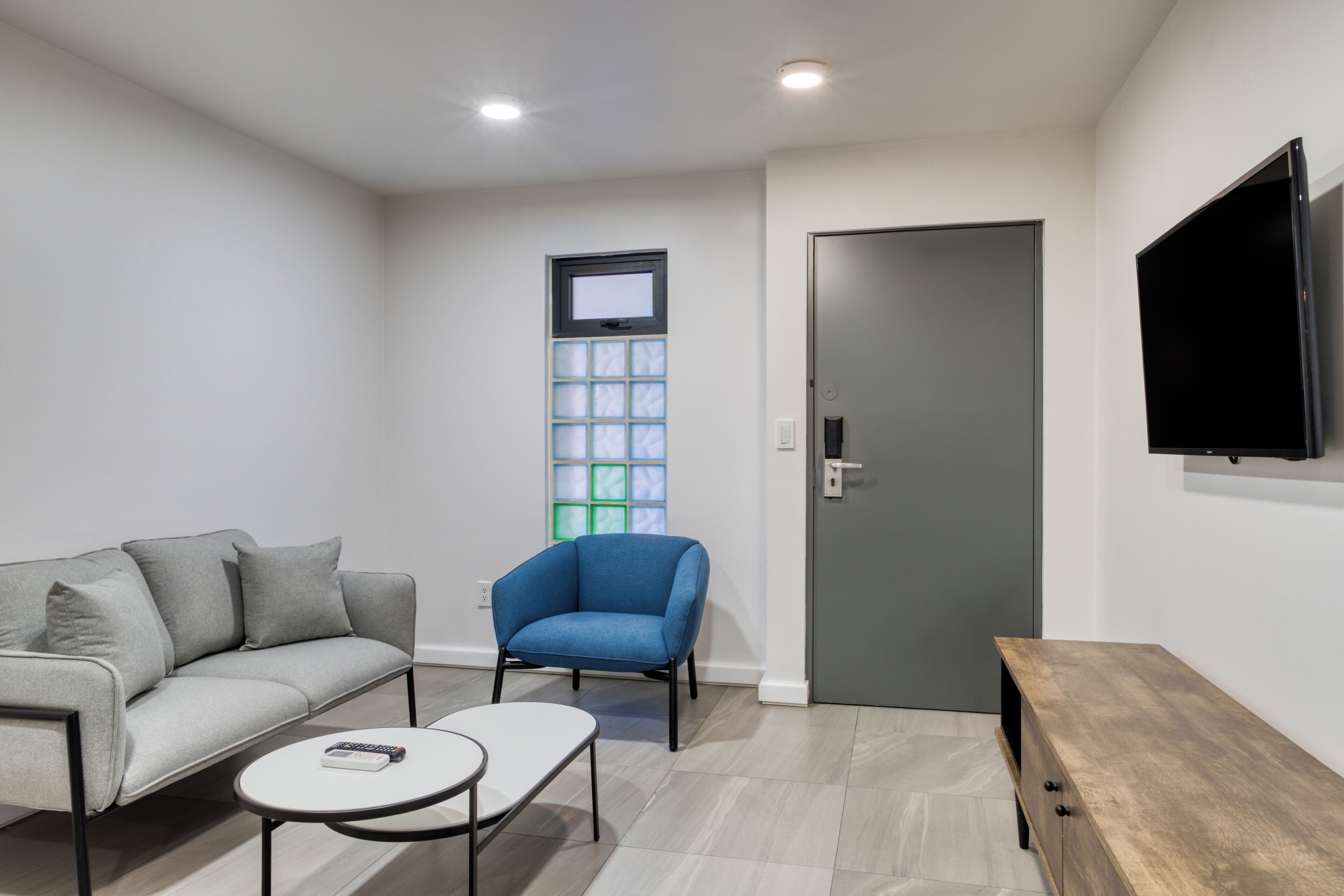 Ein modernes Wohnzimmer mit einer Couch, einem blauen Sessel, einem kleinen runden Tisch, einer Holzbank, einem an der Wand befestigten Fernseher und einer Tür mit Fenster darüber.