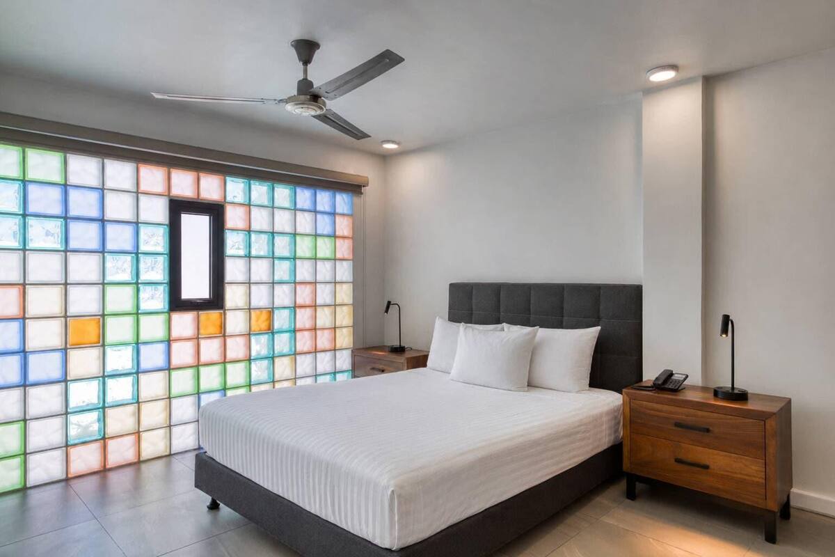 A modern bedroom with a large bed, a ceiling fan, bedside lamps, and a stained glass window.