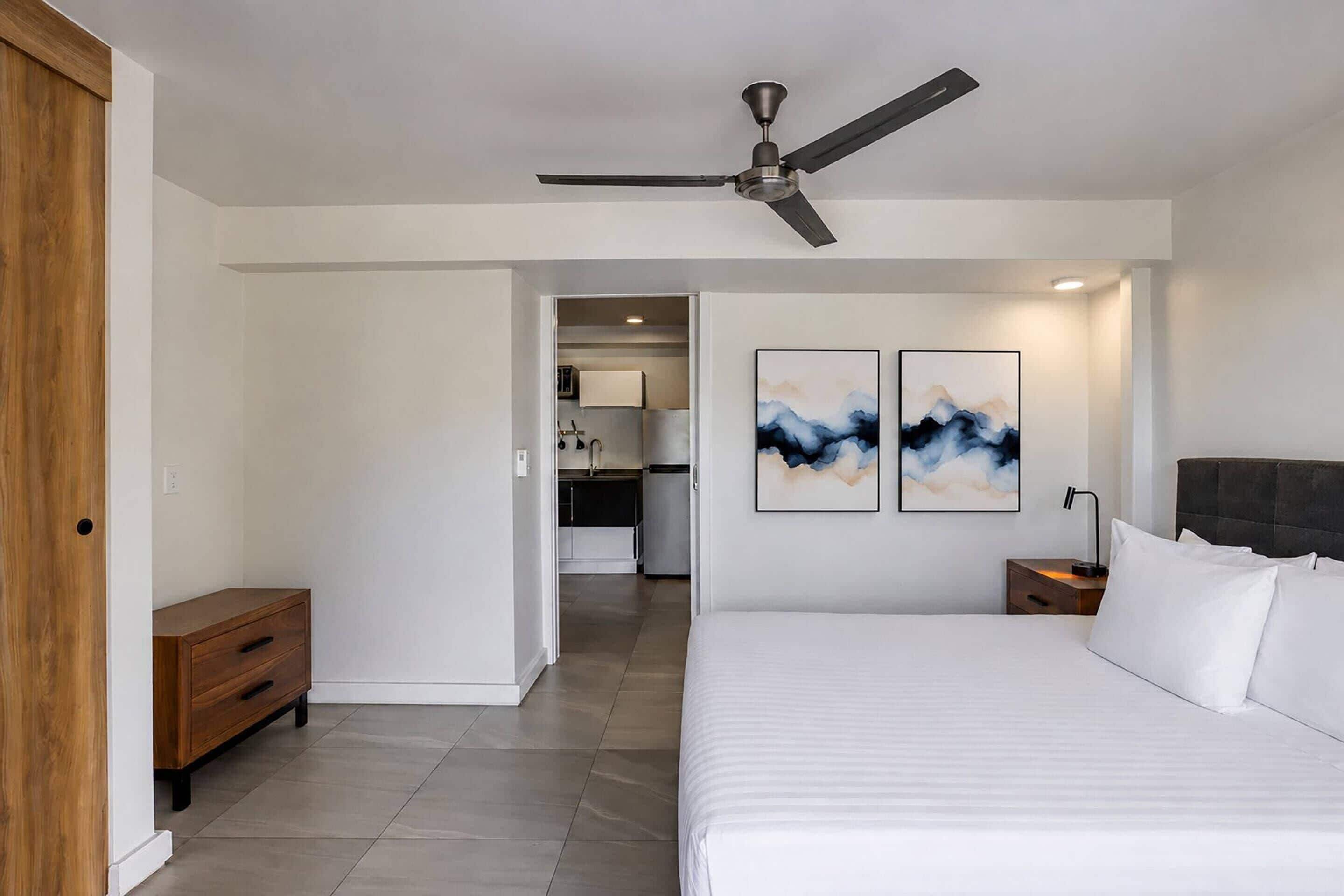 Un dormitorio con cama, mesita de noche, ventilador de techo y una obra de arte en la pared.