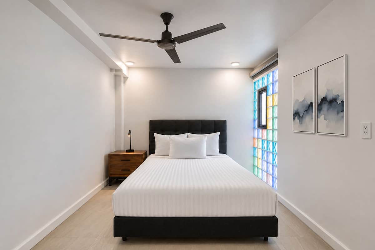 A bedroom with a bed, a nightstand, a ceiling fan, and a window with stained glass.
