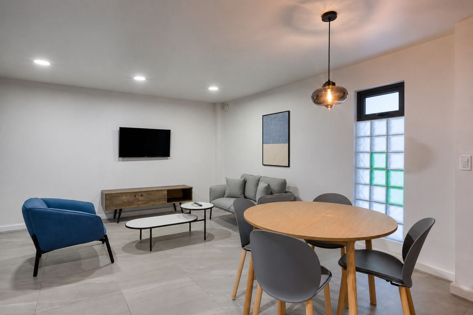 A modern living room with a round wooden table, grey sofas, a blue armchair, a wooden bench, and a TV mounted on the wall.