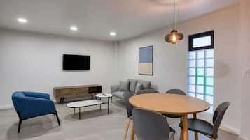 A modern living room with a round wooden table, grey sofas, a blue armchair, a wooden bench, and a TV mounted on the wall.