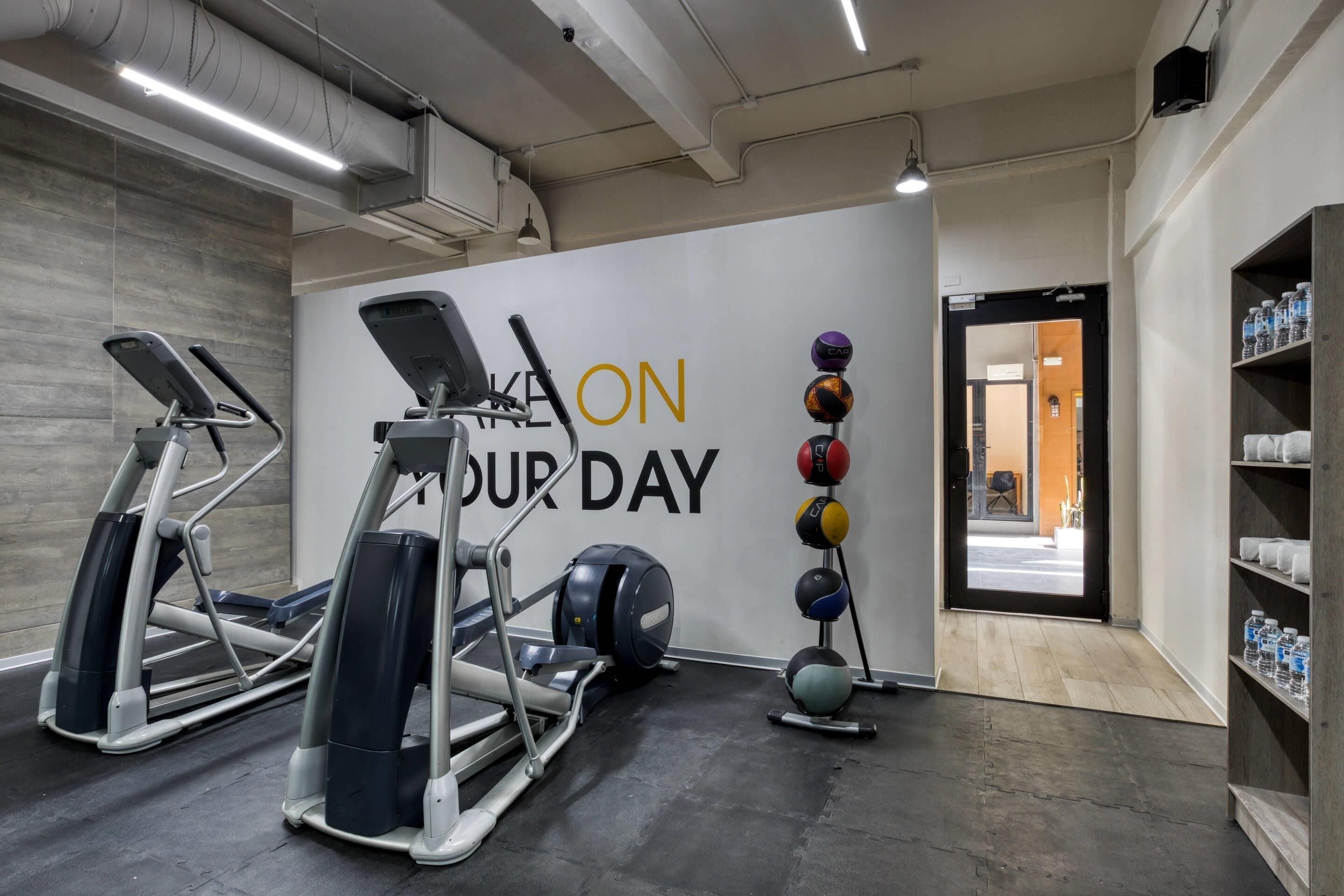 A gym with treadmills, a wall with motivational text, a rack of exercise balls, and a water bottle shelf.