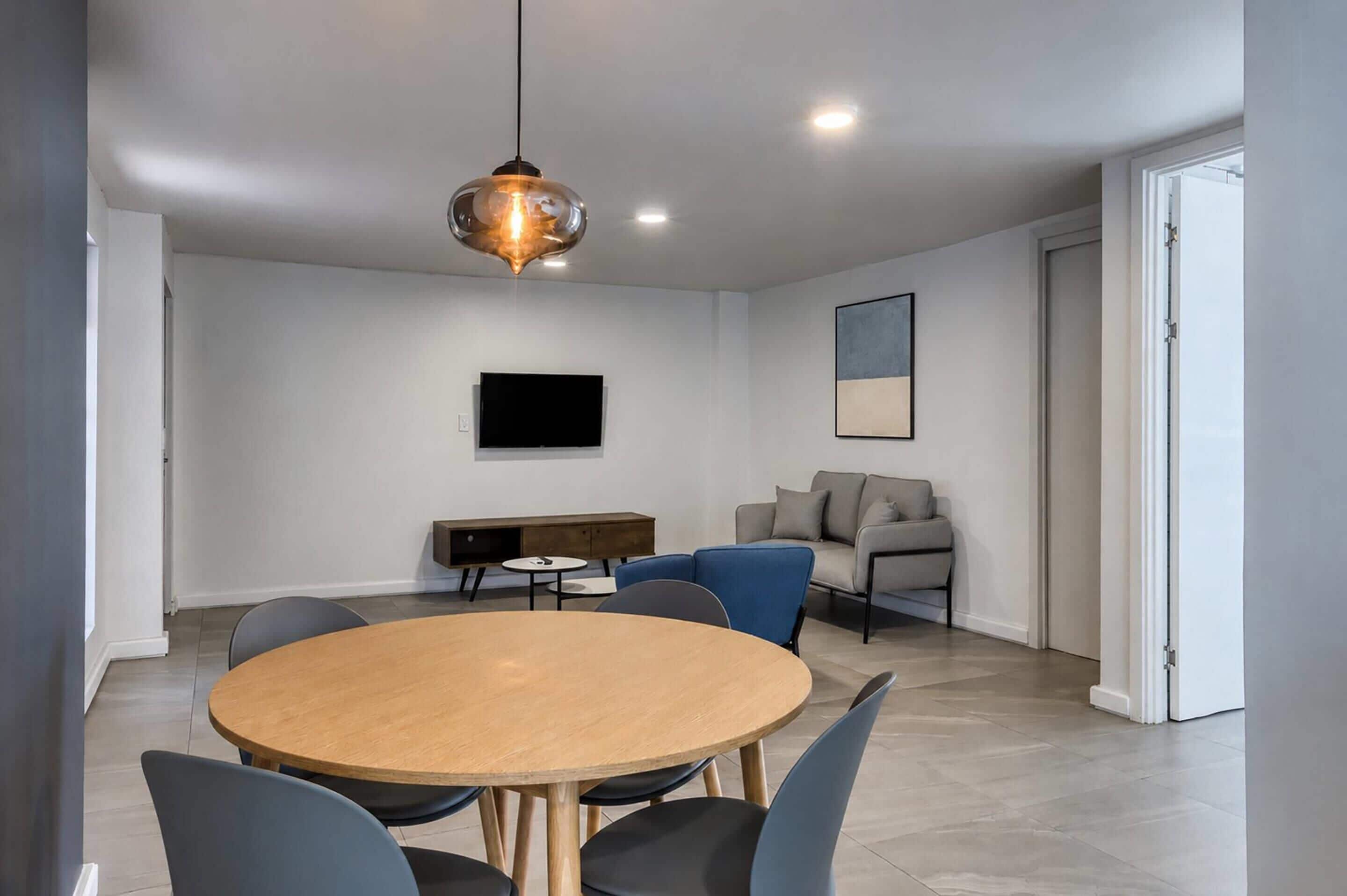 A modern living room with a round wooden table, blue chairs, a sofa, a TV mounted on the wall, and a pendant light.