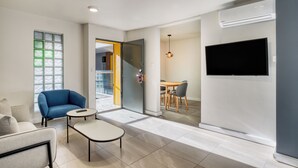 Un salon moderne avec un canapé, une petite table, un téléviseur fixé au mur et une porte vitrée donnant sur un balcon.