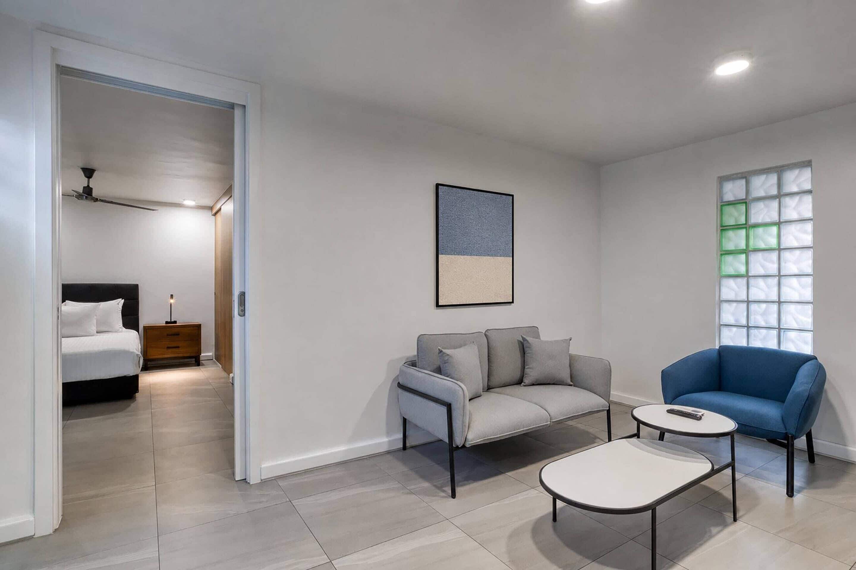 Una sala moderna con sofá, sillón y mesa de centro, una cama en la habitación contigua, y un cuadro enmarcado en la pared.