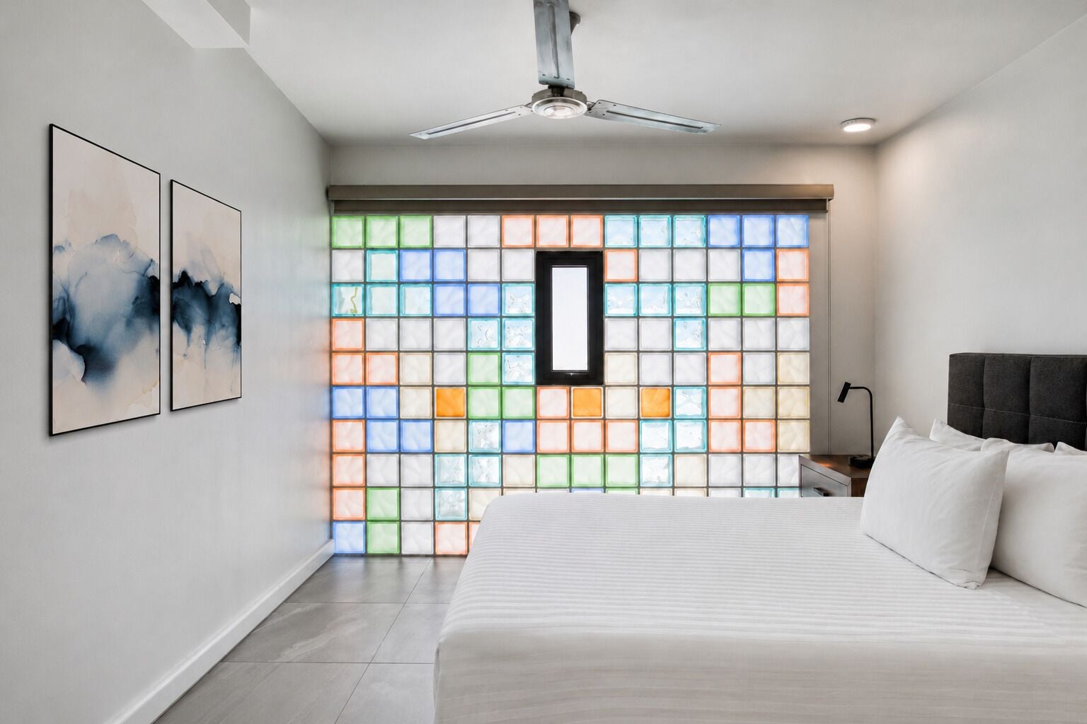A bedroom with a bed, a ceiling fan, a nightstand, and a wall with a colorful window.