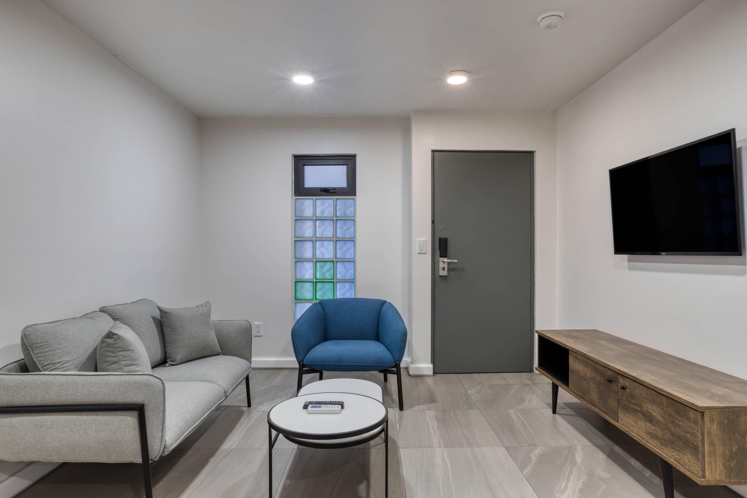A modern living room with a sofa, a blue armchair, a small round table, a wooden TV stand, and a door.
