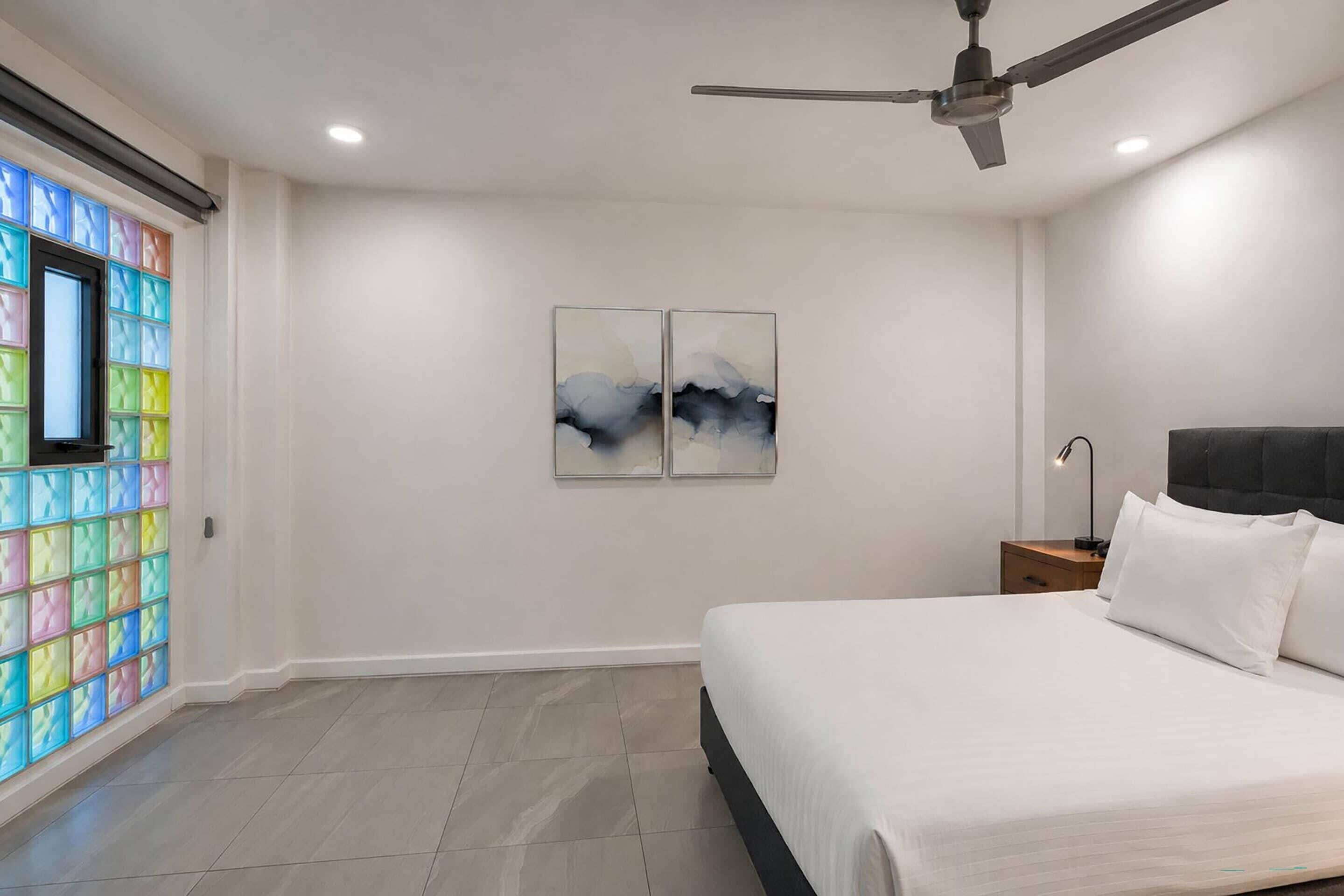 Une chambre à coucher comprenant un lit, une table de chevet, un ventilateur de plafond, des œuvres d’art et une fenêtre ornée de blocs de verre colorés.
