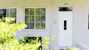 A white building with two windows and a door, a bench outside, and greenery in the foreground. - LiRi - Family paradise (Ialysos)