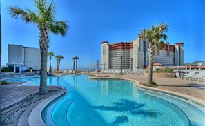 A swimming pool with palm trees, a hotel building, and lounge chairs.