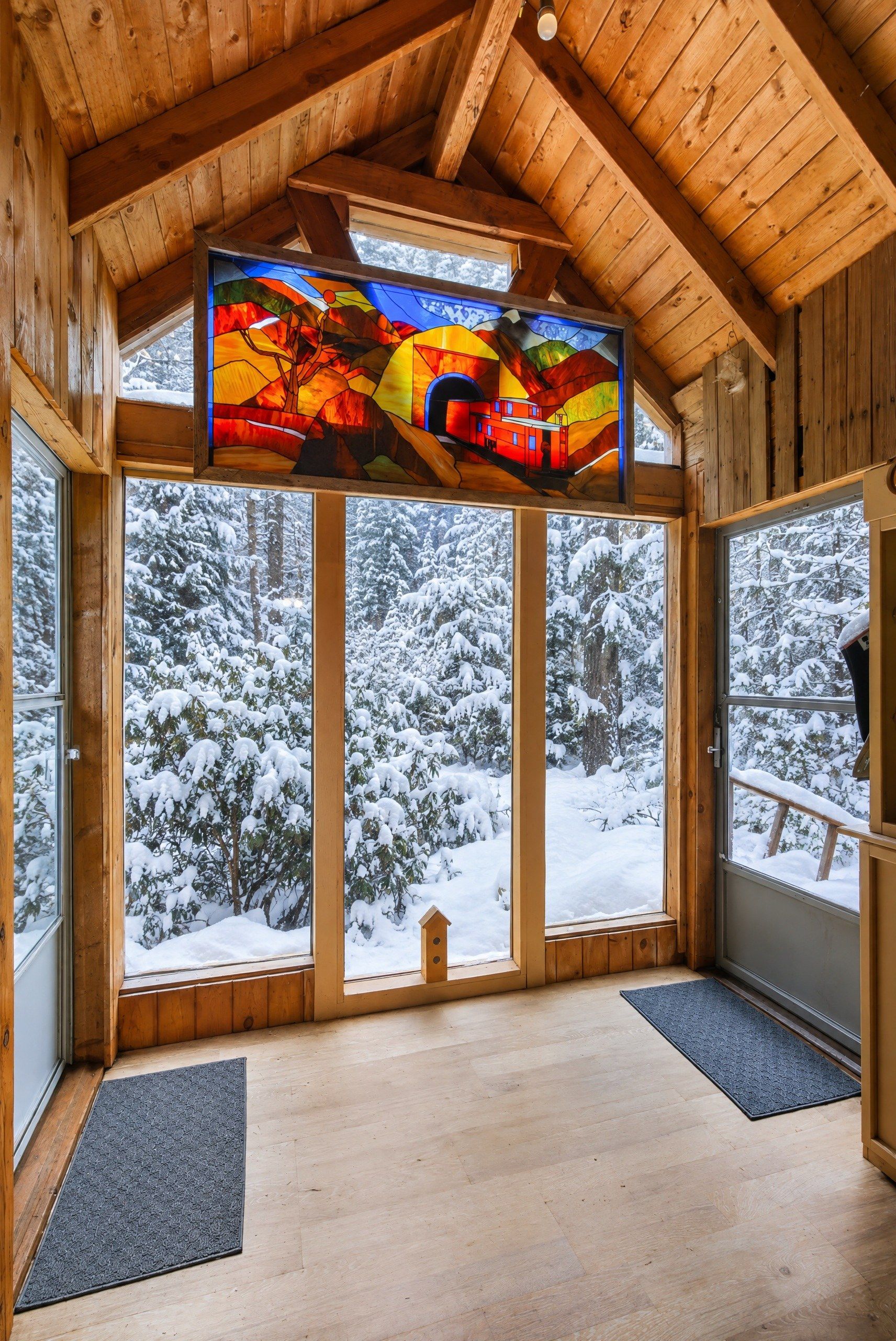 Un intérieur en bois, de grandes fenêtres, un vitrail et une vue sur des arbres couverts de neige.
