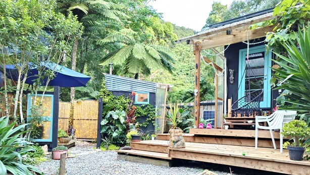 A rustic wooden structure with a porch, surrounded by lush greenery and a variety of plants.