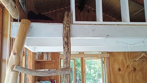 A rustic cabin interior with a loft bed, wooden walls, and a window overlooking greenery.