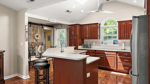 A kitchen with wooden cabinets, a stainless steel refrigerator, a bar stool, and a window with a view of greenery. - 3 Bedroom Family Retreat in Ocean Pines! (Ocean Pines)