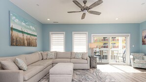 A living room with a sectional sofa, ottoman, ceiling fan, and a large painting of a beach scene.