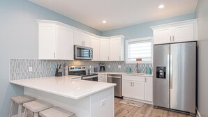 A modern kitchen with white cabinets, a stainless steel refrigerator, a microwave, and a built-in oven. There are three wooden stools and a tiled backsplash.