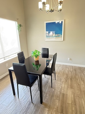 A dining area with a glass table, black chairs, a chandelier, a framed painting, and a potted plant.