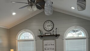 A room with a ceiling fan, two pendant lights, a clock, and a wall-mounted shelf with decorative items.