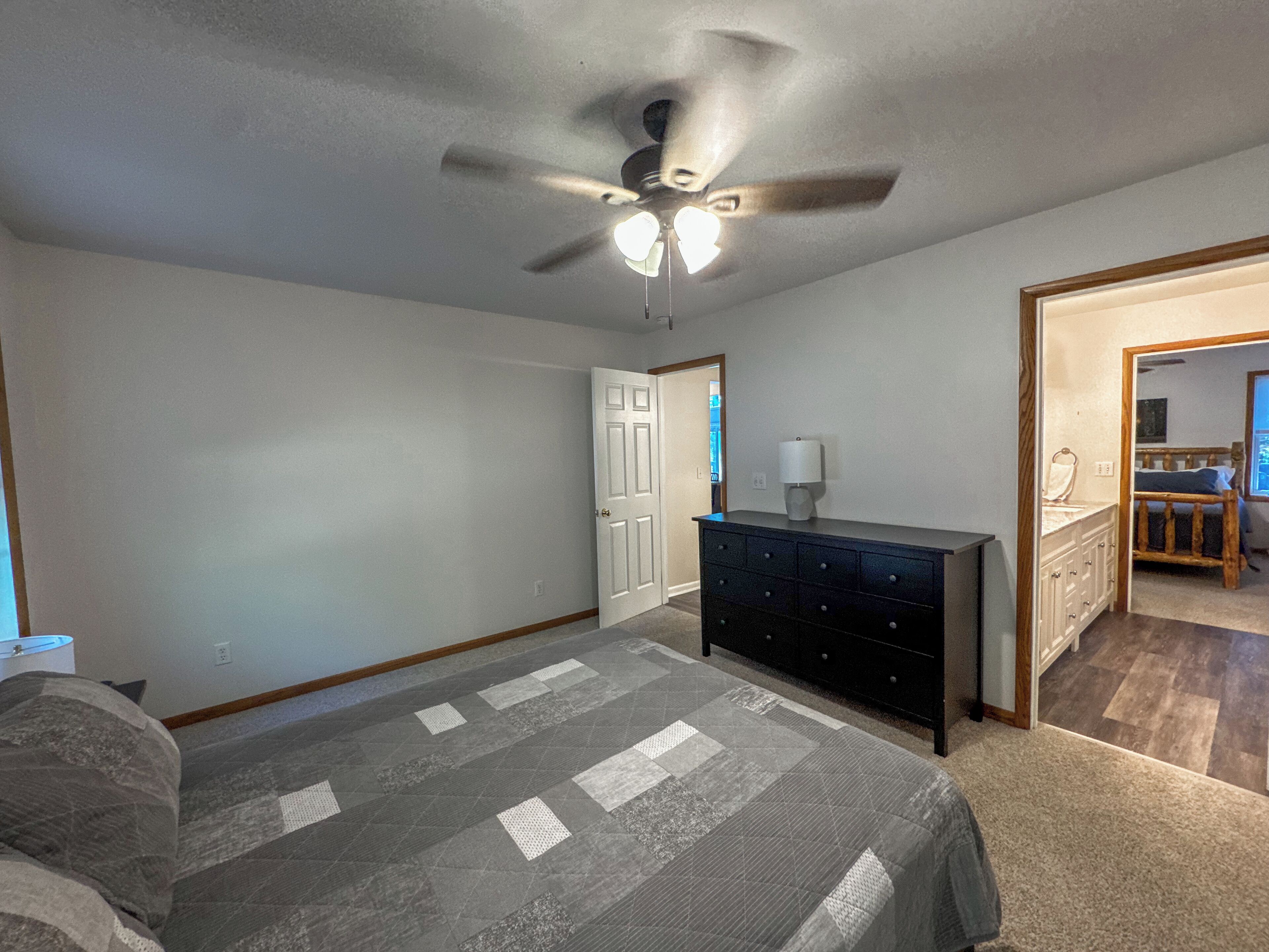 A bedroom with a bed, a dresser, a ceiling fan, and a door leading to another room.