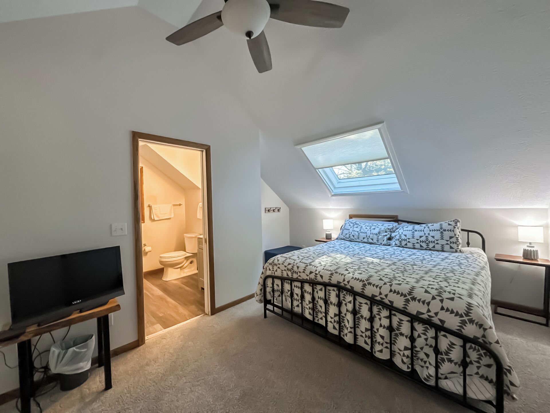 A bedroom with a bed, a TV, a ceiling fan, a skylight, and a bathroom with a toilet and sink.