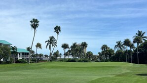 A well-maintained golf course with a clear sky, palm trees, and a building with a green roof in the background. - Ocean Village Beach Apartment on the Golf Course (Fort Pierce)
