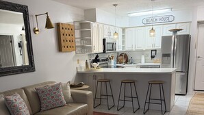 A modern kitchen with white cabinets, a central island, bar stools, and a sign reading 'BEACH HOUSE'. - Ocean Village Beach Apartment on the Golf Course (Fort Pierce)