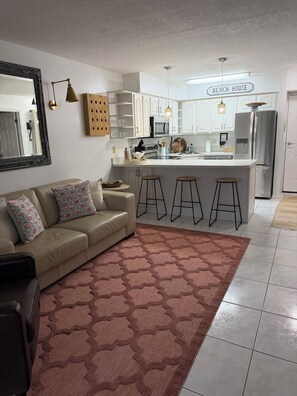 A modern kitchen with white cabinets, a central island, bar stools, and a sign reading 'BEACH HOUSE'. - Ocean Village Beach Apartment on the Golf Course (Fort Pierce)
