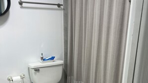 Kylpyhuone, jossa on wc, suihkuverho, kaide, roskakori ja puinen matto.
