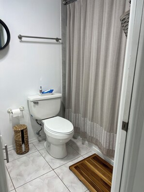 Une salle de bain équipée d’un WC, d’un rideau de douche, d’une barre d’appui, d’une poubelle et d’un tapis en bois.