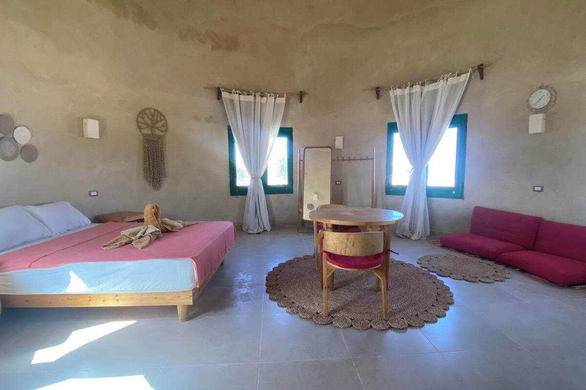 Una habitación con una cama, una mesita, un sofá rojo y dos ventanas con cortinas.