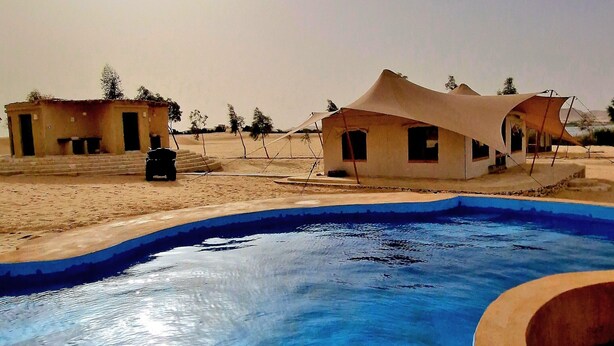 Un espace piscine agrémenté d’un bassin à l’eau d’un bleu cristallin, entouré d’un paysage aride évoquant le désert et de bâtiments simples, de plain-pied.