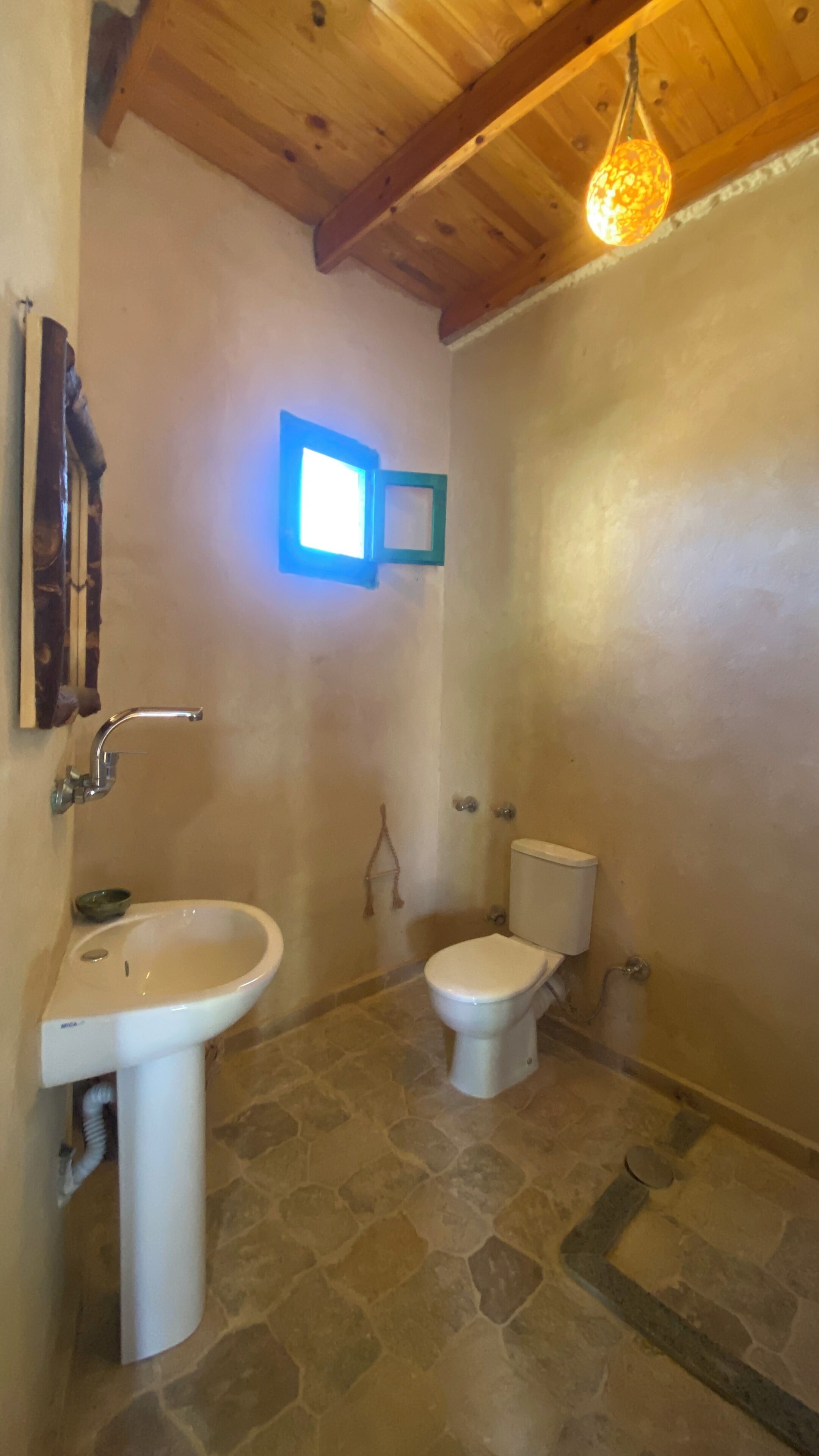 Ein Badezimmer mit Steinboden, einem weißen Waschbecken, einer Toilette und einer Holzbalkendecke.