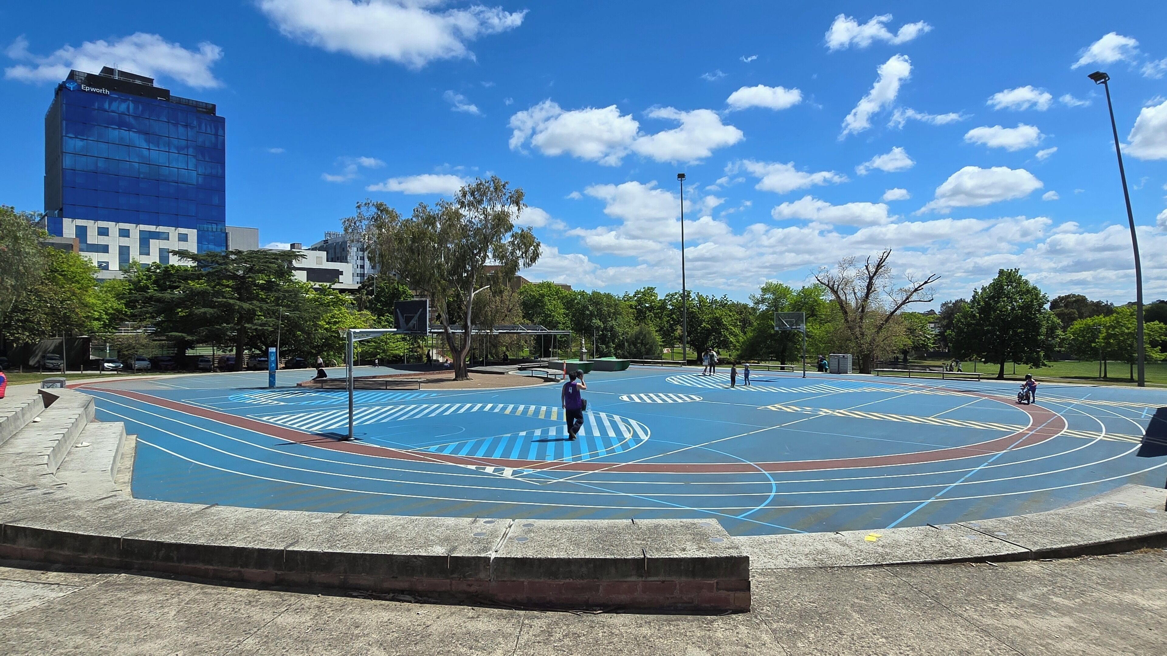 Una cancha de baloncesto al aire libre con pista azul y roja, rodeada de árboles y un edificio moderno al fondo.