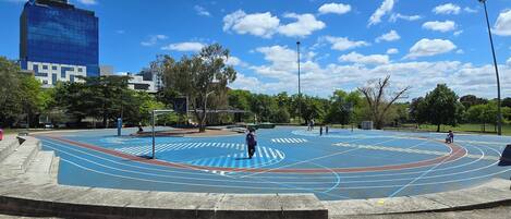 Una cancha de baloncesto al aire libre con pista azul y roja, rodeada de árboles y un edificio moderno al fondo.