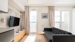 Un salon moderne avec un canapé gris, un téléviseur à écran plat fixé au mur, une petite table ronde et une vue sur la ville grâce à de grandes fenêtres.