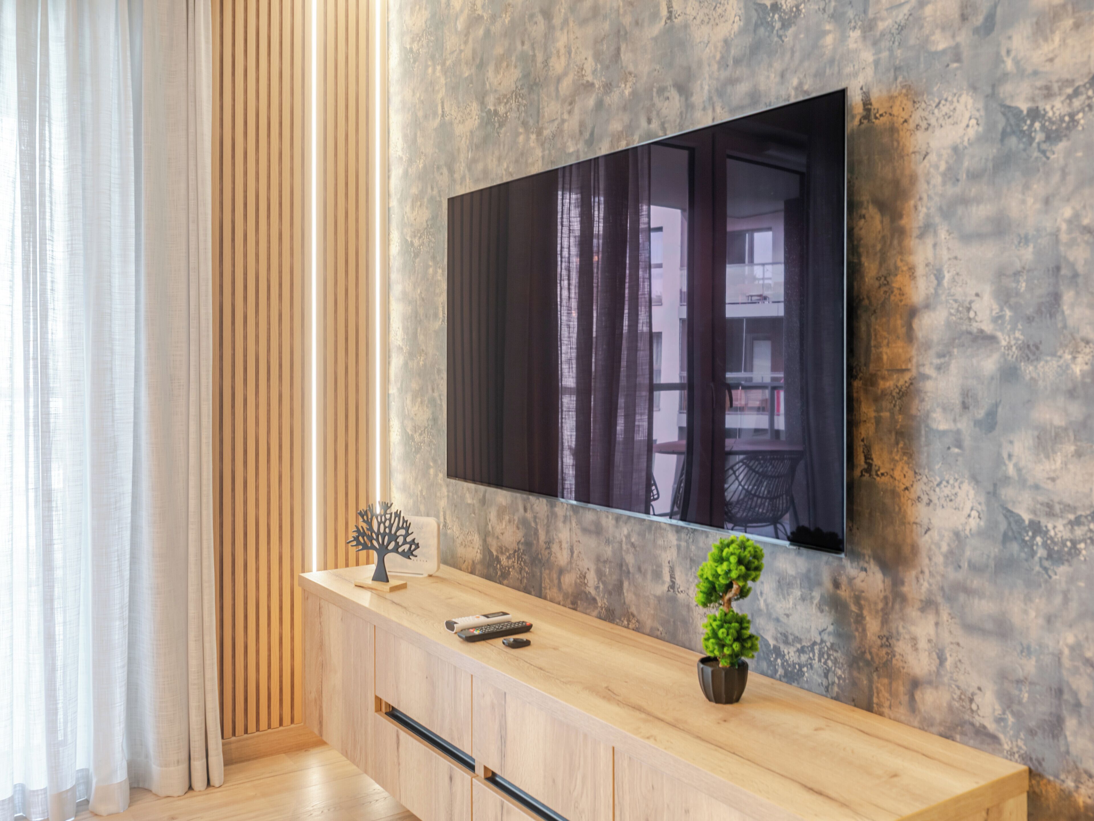 Un soggiorno moderno con un tavolo consolle in legno, una TV a schermo piatto montata su una parete strutturata, una piccola pianta in vaso e una scultura arborea decorativa.