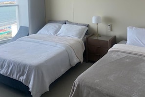 Habitación de hotel con dos camas, una mesita de noche con lámpara y vista a la playa.