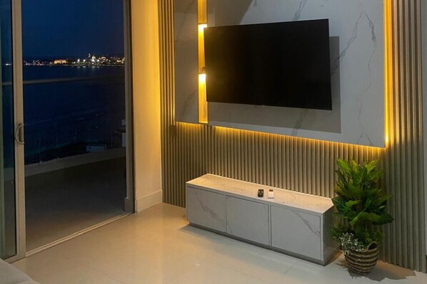 Una sala de estar moderna con un televisor grande montado en la pared, un ventanal que da a un balcón y un mueble bajo minimalista con una planta.