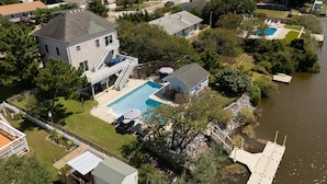 An aerial view of a residential area with a large house featuring a two-story deck, a swimming pool, and a dock extending into a body of water.
