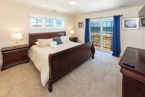 A bedroom with a large bed, two bedside tables, a nightstand, a dresser, a window with a view of the beach, and a balcony with blue curtains. - Bellissimo - Oceanfront with Pool, Hot Tub, Theater Room, Elevator, Game Tables (Virginia Beach)