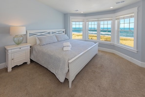A bedroom with a bed, a nightstand, a lamp, and a view of the beach. - Bellissimo - Oceanfront with Pool, Hot Tub, Theater Room, Elevator, Game Tables (Virginia Beach)