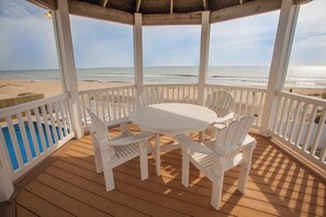 A covered outdoor area with a wooden deck, white chairs, and a table, overlooking a beach and ocean. - Bellissimo - Oceanfront with Pool, Hot Tub, Theater Room, Elevator, Game Tables (Virginia Beach)