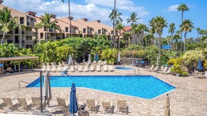 A resort pool area with lounge chairs and umbrellas, surrounded by palm trees and a building with balconies. - Charming Beachside Garden Vistas (Kihei)
