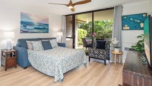 A bedroom with a bed, a ceiling fan, a nightstand, a chair, a desk, and a large window with a view of greenery. - Charming Beachside Garden Vistas (Kihei)