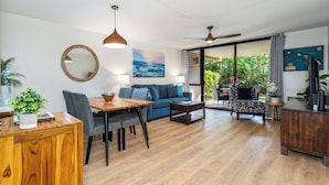 A modern living room with a dining area, a sofa, a coffee table, and a TV. There is a large window with a view of greenery and a painting on the wall. - Charming Beachside Garden Vistas (Kihei)